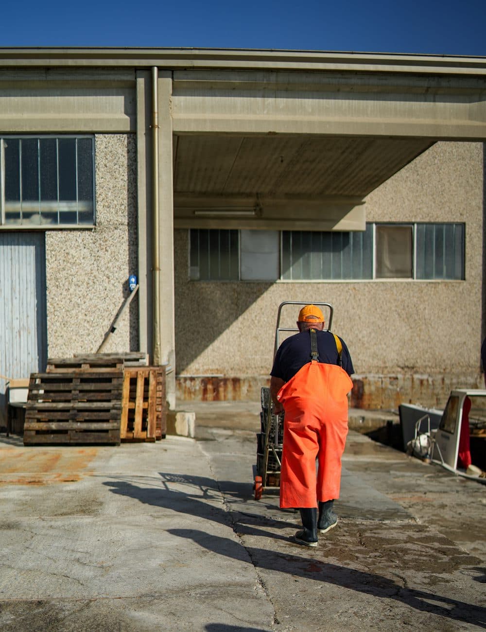 A worker in orange overalls pushing a cart across a yard